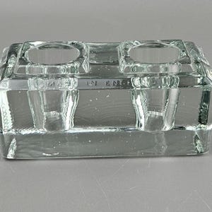 Peut inclure: Un encrier en verre transparent avec deux puits circulaires et deux porte-plumes. L'encrier est rectangulaire avec un dessus et une base plats. Les mots "MADE IN ENGLAND" sont gravés sur le devant.