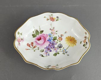 May include: A white, scalloped-edge dish with a gold rim, adorned with a floral design. The flowers include a large pink rose, blue, purple, and yellow blossoms, and green leaves. A decorative piece for serving or display.