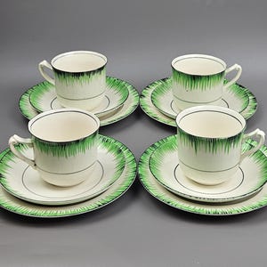Puede incluir: Cuatro tazas de té y platillos a juego. Las tazas y los platillos son blancos con un diseño verde y negro. Las tazas tienen un asa y los platillos tienen un borde verde y negro a juego. El juego está dispuesto sobre una superficie gris.