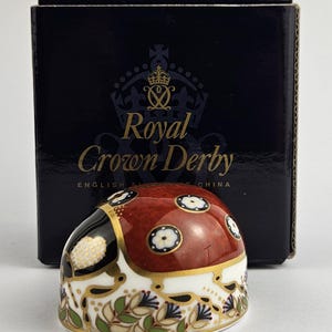 Peut inclure: Un presse-papier en porcelaine en forme de coccinelle avec un dos rouge orné de motifs floraux blancs et bleus, une tête noire et une base blanche décorée de feuilles vertes et bleues. Derrière se trouve une boîte bleue avec Royal Crown Derby.