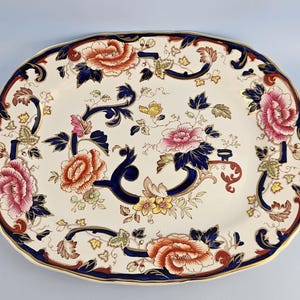 May include: An oval ceramic serving platter with a cream-colored base featuring a decorative pattern of pink, orange, and yellow flowers, green leaves, and dark blue scrolling accents. The scalloped rim is trimmed with a thin gold-colored line.