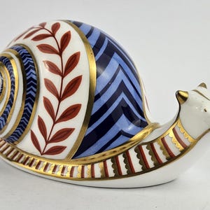 Può includere: Statuina decorativa a forma di lumaca in ceramica. Il guscio presenta un motivo a spirale, un motivo a zig-zag blu e accenti di foglie rosse, il tutto bordato in oro. Il corpo della lumaca è bianco con finiture dorate.