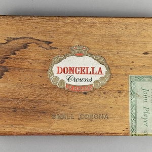 May include: Wooden cigar box with the text "DONCELLA Crowns" in red and white on a decorative label. The box has a distressed appearance with a faded green label on the right side. The text "Still Corona" is visible.