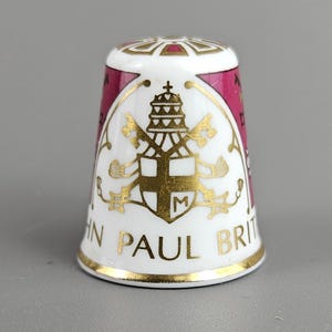 Puede incluir: Un dedal de cerámica blanco con detalles dorados y burdeos. El dedal presenta un emblema dorado y el texto "JOHN PAUL BRIT". La parte superior tiene un diseño geométrico dorado y burdeos.