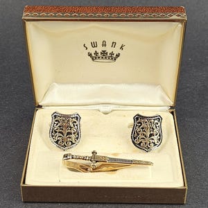 Puede incluir: Un conjunto de joyería Swank en una caja marrón que incluye dos gemelos en forma de escudo con filigrana dorada sobre fondo negro y un pasador de corbata dorado con forma de espada con grabados en la empuñadura y la hoja.