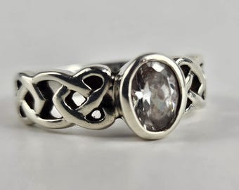 Könnte beinhalten: Ein silberner Ring mit einem keltischen Knotenmuster auf dem Band und einem ovalen, klaren Edelstein. Der Ring ist vor einem weißen Hintergrund platziert, der die komplizierten Details der Metallarbeiten und das Funkeln des Steins hervorhebt.