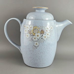 Op de afbeelding: Lichtblauwe keramische theepot met een deksel. De theepot heeft een bloemenpatroon van witte en gele bloemen. De handgreep en de tuit zijn gebogen. De theepot heeft een gespikkelde textuur.