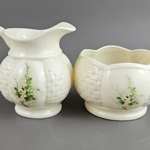Puede incluir: Juego de jarra y azucarero de cerámica color crema. La jarra tiene asa y pico, mientras que el cuenco tiene un borde festoneado. Ambas piezas presentan un diseño de diamantes y floral con detalles en verde y blanco.