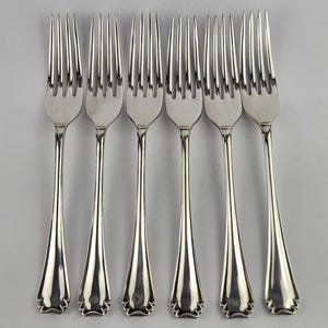 Puede incluir: Seis tenedores plateados dispuestos en fila. Cada tenedor tiene cuatro púas y un mango largo y delgado. Los mangos tienen un diseño decorativo festoneado en la base. Los tenedores son de metal brillante.