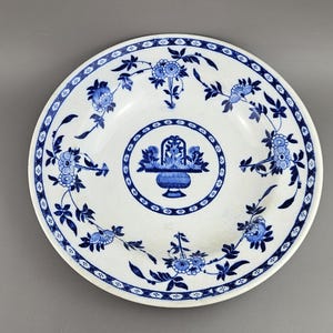 May include: White ceramic plate with a blue floral design. The plate features a central floral basket motif and a repeating floral pattern around the rim. The plate's design is in a classic, vintage style.