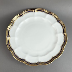 May include: White scalloped-edge plate with a gold and navy blue border. The border features a gold leaf design and a navy blue band with small gold dots. A decorative piece for serving food.