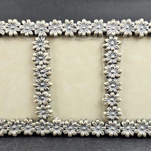 May include: A silver-toned triple photo frame with a daisy flower design. The frame has three rectangular openings for photos and is decorated with small daisy flowers around the edges. The frame is made of metal and has a vintage appearance.