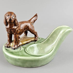 Puede incluir: Un cenicero de cerámica vintage con una figura de perro marrón. El perro está de pie sobre una pequeña base de color canela, colocada sobre un cenicero verde claro en forma de hoja con un reposo curvo. El cenicero tiene un acabado brillante.