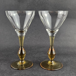 May include: Two vintage clear glass cocktail glasses with amber-colored stems and bases. The glasses have a classic V-shaped bowl design, perfect for serving cocktails. The stems have a decorative, bulbous shape.