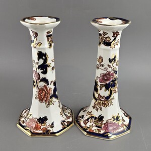 May include: A pair of white ceramic candlesticks featuring floral and foliage designs in navy, gold, and red-pink hues. The candlesticks have a hexagonal base and a flared form, with a height of approximately 20 cm.
