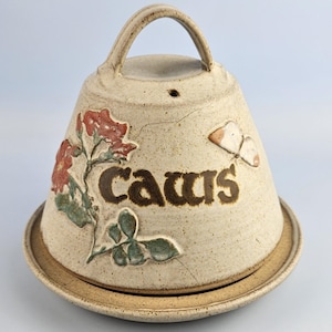May include: A beige speckled ceramic butter bell with a handle on top. The exterior features a raised relief design of red flowers with green leaves and a small butterfly. The word "caws" is embossed in brown lettering on the front of the bell.