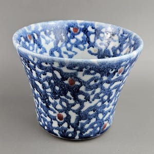 Può includere: Una tazza in ceramica dalla forma svasata, con un motivo blu e bianco con piccoli accenti rossi. Il design copre l'esterno della tazza, creando un aspetto strutturato. Il bordo è una tonalità più scura di blu.