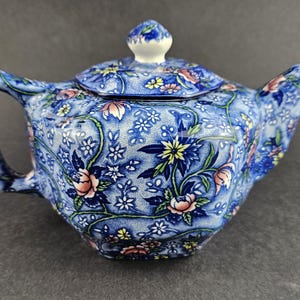 May include: A ceramic teapot featuring a blue and white floral pattern with accents of pink and yellow. The teapot has a matching lid with a small knob, a curved handle, and a spout. The surface is covered in a dense, intricate botanical design.