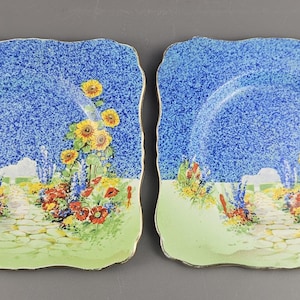 Könnte beinhalten: Zwei quadratische Teller mit blauem, gesprenkeltem Hintergrund und einer Gartenszene. Die Teller haben einen hellgrünen Rand und einen goldenen Rand. Die Gartenszene umfasst Sonnenblumen, einen Steinweg und bunte Blumen.