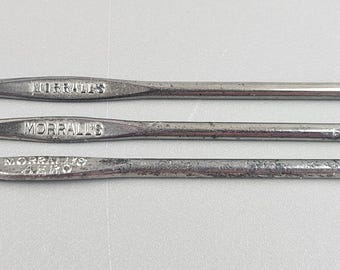 Op de afbeelding: Drie vintage zilveren haaknaalden van verschillende maten, horizontaal gerangschikt. Elke haaknaald heeft een slanke, langwerpige handgreep met reliëftekst. De haaknaalden zijn van metaal en staan tegen een effen, lichtgekleurde achtergrond.