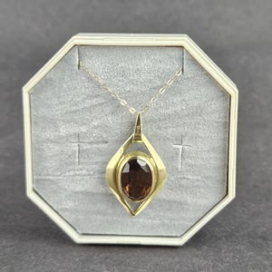 Könnte beinhalten: Eine goldene Anhängerkette mit einem oval geschliffenen, dunkelbraunen Edelstein in einem geometrischen, rautenförmigen Rahmen. Der Anhänger hängt an einer feinen Goldkette und befindet sich in einer achteckigen Schatulle mit grauem Samtfutter.