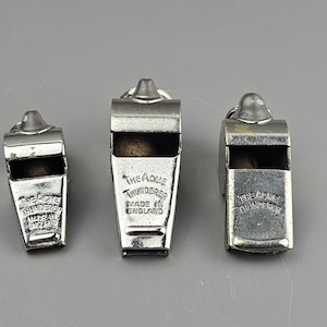 May include: Three silver-toned Acme Thunderer whistles of varying sizes. The whistles have a rectangular body with a rounded top and a ring for attachment. The text "THE ACME THUNDERER MADE IN ENGLAND" is visible on the front of the whistles.
