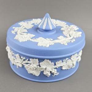 May include: A round, light blue trinket box with a white, raised relief design of grape vines and leaves. The lid has a conical finial and the box is approximately 4 inches in diameter.