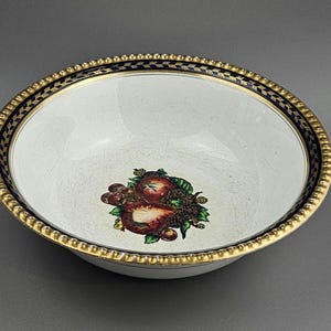 Può includere: Ciotola in ceramica bianca con bordo dorato perlato e bordo a motivi blu e oro. La ciotola presenta un disegno di frutta al centro, tra cui fragole e uva.