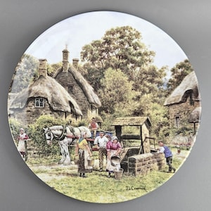 May include: A decorative plate featuring a rural village scene. The image depicts thatched-roof cottages, a horse-drawn cart, and people carrying buckets. The plate has a glossy finish and a detailed, colorful illustration of a pastoral setting.