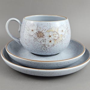 Op de afbeelding: Een lichtblauwe keramische theekop en schotel set. De kop heeft een afgeronde vorm met een handvat en is versierd met een bloemenpatroon. De set bevat twee bijpassende schotels met een bruine rand.