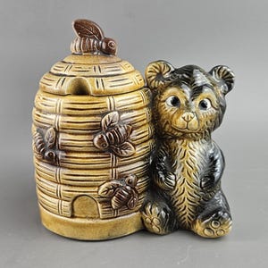 May include: A ceramic honey pot and bear figurine. The honey pot is shaped like a beehive with a lid and a bee on top. The bear is brown and black, sitting next to the beehive. The pot has bee and leaf details.