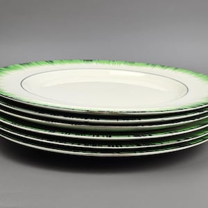 Puede incluir: Una pila de seis platos de cerámica blancos. Cada plato tiene un diseño verde, similar a un pincel, alrededor del borde y una fina línea negra cerca del borde del centro blanco. Los platos están apilados uno encima del otro.
