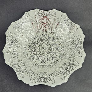 Op de afbeelding: Een decoratieve witte schaal met een zilveren bloemenpatroon. De schaal heeft een geschulpte rand en een gedetailleerd, symmetrisch patroon in het midden. Het ontwerp bevat bloemen, krullen en een stervorm.