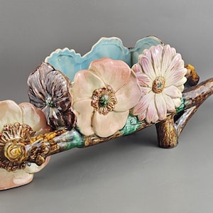 Puede incluir: Centro de mesa floral de cerámica con forma de carretilla. La pieza presenta grandes flores detalladas en tonos rosas, blancos y marrones, con un interior azul claro. La carretilla es marrón con detalles verdes.