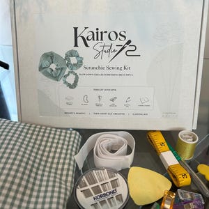 Puede incluir: Un kit de costura de coleteros con el texto "Kairos Studio 72 Scrunchie Sewing Kit". El kit incluye tela, elástico, agujas, hilo, una cinta métrica y otros suministros de costura. La tela tiene un estampado de cuadros verdes y blancos.