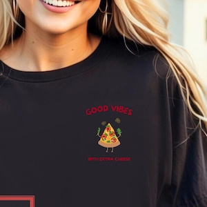 Könnte beinhalten: Schwarzes T-Shirt mit einem Pizzastück-Motiv und dem Text "Good Vibes With Extra Cheese". Das Pizzastück hat ein lächelndes Gesicht und Arme. Das gleiche Design wiederholt sich kleiner auf der rechten Brust.