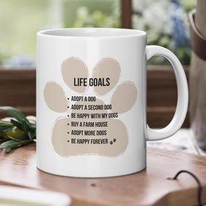 Könnte beinhalten: Eine weiße Keramiktasse mit einem braunen Pfotenabdruck-Design. Die Tasse zeigt den Text "LIFE GOALS" und eine Liste von hundefreundlichen Zielen, darunter "Einen Hund adoptieren", "Einen zweiten Hund adoptieren", "Mit meinen Hunden glücklich sein", "Ein Bauernhaus kaufen", "Mehr Hunde adoptieren" und "Für immer glücklich sein".