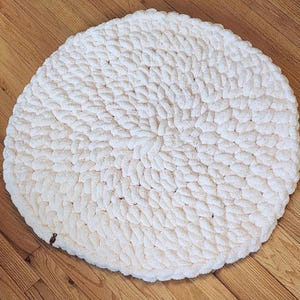 May include: A white round knitted rug with a textured surface. The rug is made of thick yarn and has a circular shape.