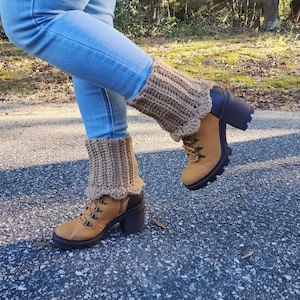 May include: Tan crochet leg warmers with a scalloped edge are worn over brown and tan hiking boots with black heels. The boots have brown laces and metal eyelets. The person is wearing blue jeans.