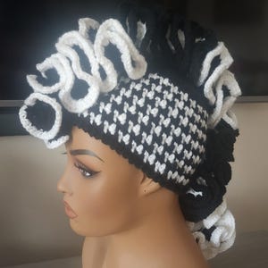 May include: A black and white crochet hat with a unique design. The hat features a houndstooth pattern on the main body and is adorned with white, ruffled trim. The hat is displayed on a mannequin head.