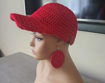 May include: A red crocheted hat with a visor and matching red crocheted earrings. The hat and earrings are made of a textured yarn. The hat has a small metal accent on the side. The earrings are large, round, and dangle from silver hooks.