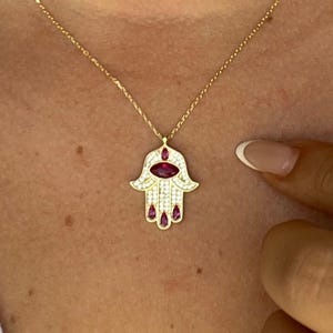 May include: Gold-toned necklace featuring a Hamsa hand pendant. The pendant is adorned with a large oval ruby in the center and smaller ruby accents. The hand is outlined with small, clear gemstones. The necklace is worn on a person.