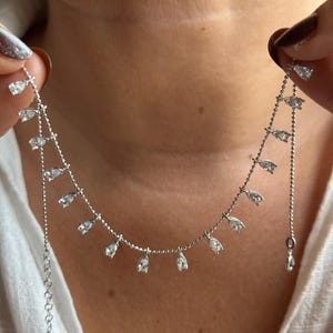 以下が含まれることがあります： 一連のしずく型の透明な宝石をあしらった繊細なシルバーネックレス。 ネックレスは、前面に均等に配置された宝石が付いた細いチェーンが付いています。 ネックレスは白いシャツに対して表示されています。