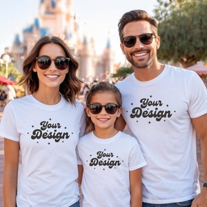 以下が含まれることがあります： レトロなフォントで「Your Design」と書かれた白いTシャツを着た3人家族。Tシャツにはテキストの周りに小さな星が散りばめられています。家族は城のような建物の前に立っています。