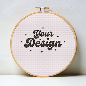 May include: A white embroidery hoop with a pink fabric background. The fabric has the text "Your Design" in black with a retro font and small stars around the text.