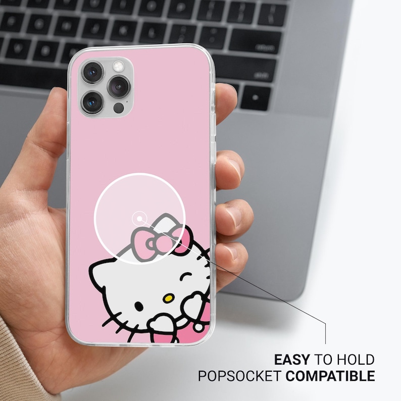May include: Pink phone case featuring a Hello Kitty design. The case has a clear popsocket circle and the text "EASY TO HOLD POPSOCKET COMPATIBLE". The phone case is held in a person's hand.