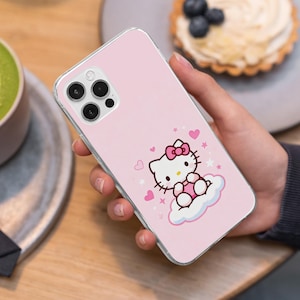 May include: Pink phone case featuring a Hello Kitty design. The case has a clear border and a light pink background. The Hello Kitty graphic includes a pink bow, hearts, and stars. The phone case is held in a person's hand.