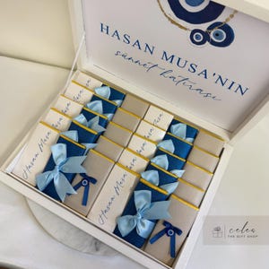 May include: A white box with a blue and gold lid. The lid has the text "Hasan Musa'nin sünnet hatırası" and a blue evil eye charm. The box is filled with small white and blue gift boxes tied with blue and white ribbons.