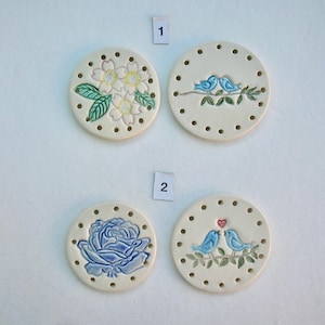 May include: Four off-white ceramic coasters with decorative designs. One features white flowers, another two blue birds on a branch, a third a blue rose, and the last two birds with a heart. Each coaster has small holes around the edge.