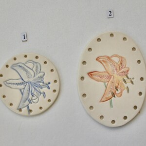 May include: Two off-white ceramic decorative pieces. One is round with a blue floral design, the other is oval with an orange floral design. Both have small holes around the edges.